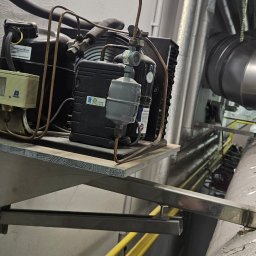 Hochlik sp. z o.o. - Zamontowany kompresor klimatyzacji na metalowej półce w pomieszczeniu technicznym, widoczne rury i instalacje. Szare ściany i sufit.