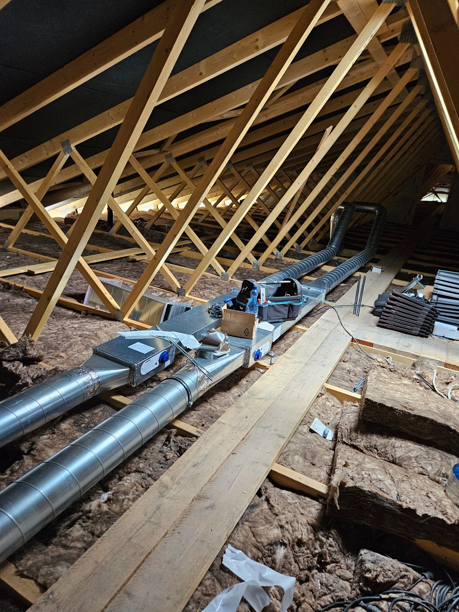 Dwie metalowe rury wentylacyjne w przestrzeni poddasza, prowadzone między drewnianymi belkami stropowymi, otoczone izolacją z wełny mineralnej. Widoczne narzędzia i materiały montażowe.