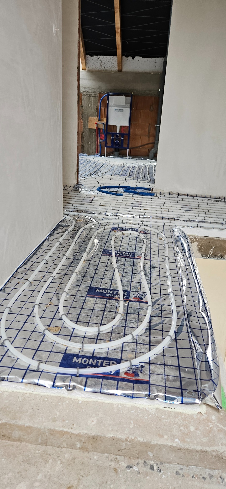 Białe rury ogrzewania podłogowego ułożone na folii z nadrukiem MONTER, widoczne elementy instalacji sanitarnej w surowym wnętrzu.