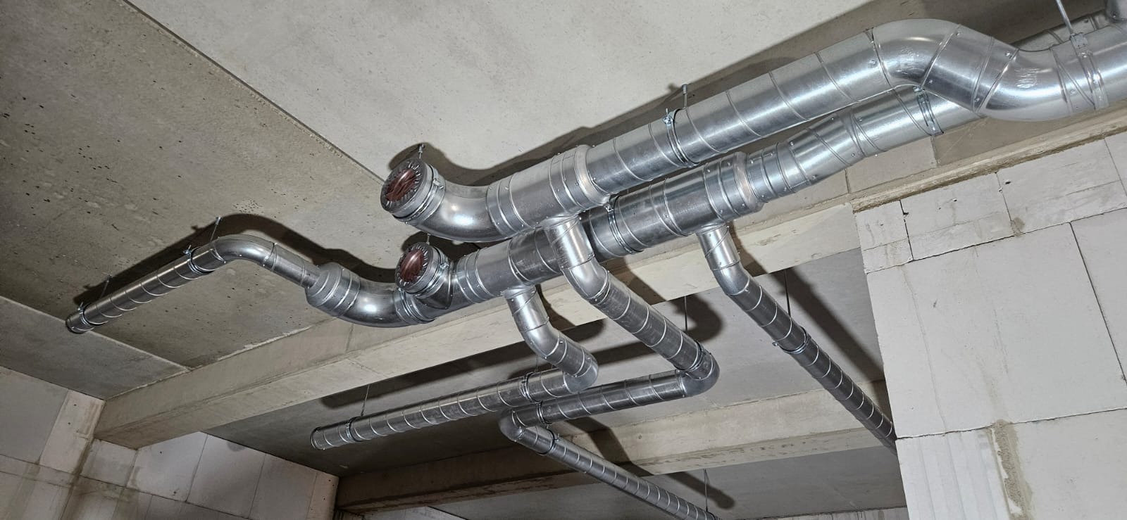 Srebrne, spiralne rury wentylacyjne zamontowane pod betonowym sufitem w surowym wnętrzu. Widoczne łączenia i mocowania.