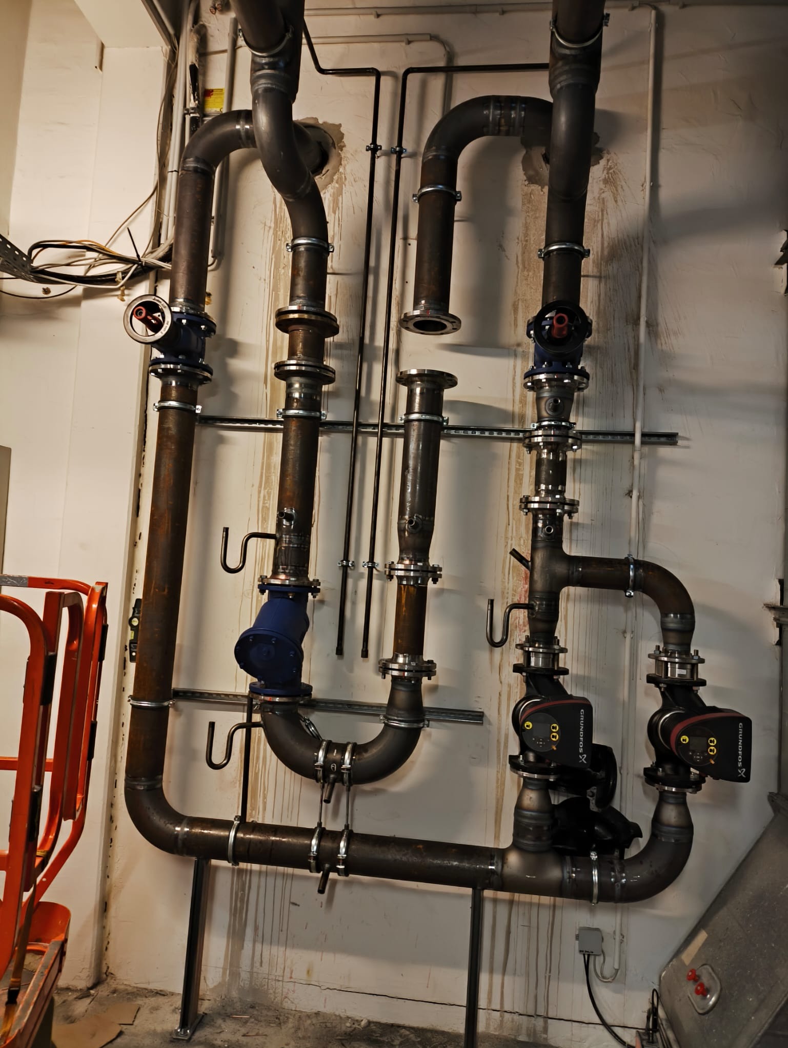 Złożona instalacja rur z zaworami i pompami Grundfos na ścianie. Widoczne spawane łączenia i elementy mocujące. Instalacja w pomieszczeniu technicznym.