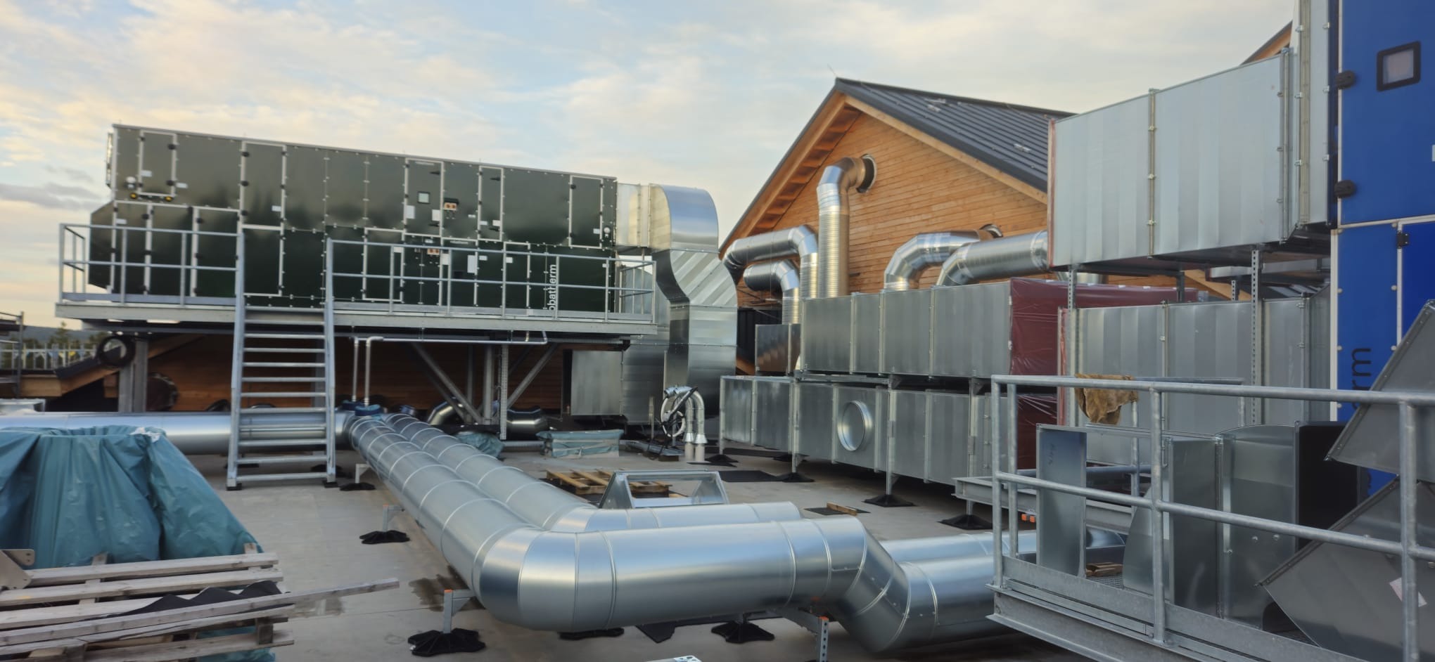 Rozbudowana instalacja wentylacyjna na dachu budynku z drewnianą konstrukcją. Widoczne liczne rury, kanały wentylacyjne i urządzenia. Elementy instalacji w kolorze srebrnym i zielonym.