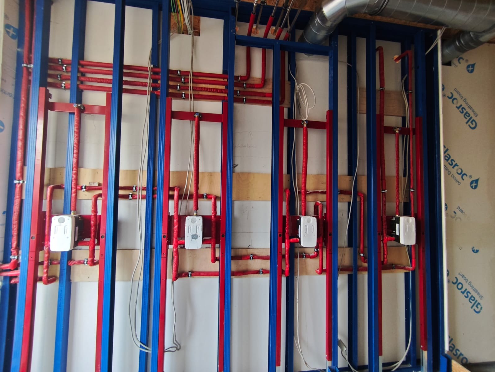Czerwone rury instalacji wodnej w izolacji, zamontowane na niebieskiej konstrukcji nośnej, z białymi puszkami przyłączeniowymi. Widoczny fragment wentylacji.