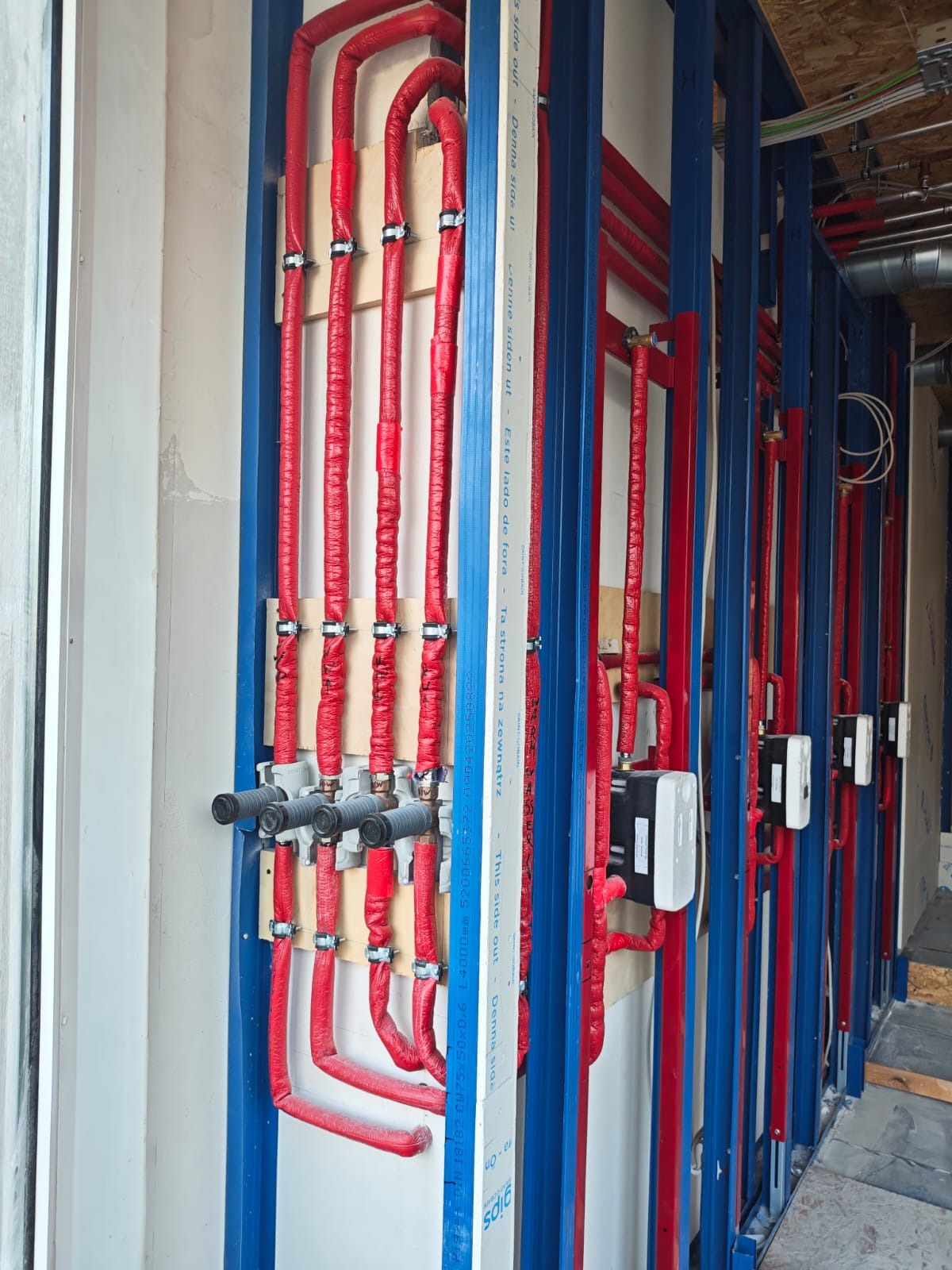 Czerwone, zaizolowane rury instalacji grzewczej zamontowane na drewnianych panelach, w konstrukcji z niebieskich profili. Widoczne czarne zawory i białe regulatory.