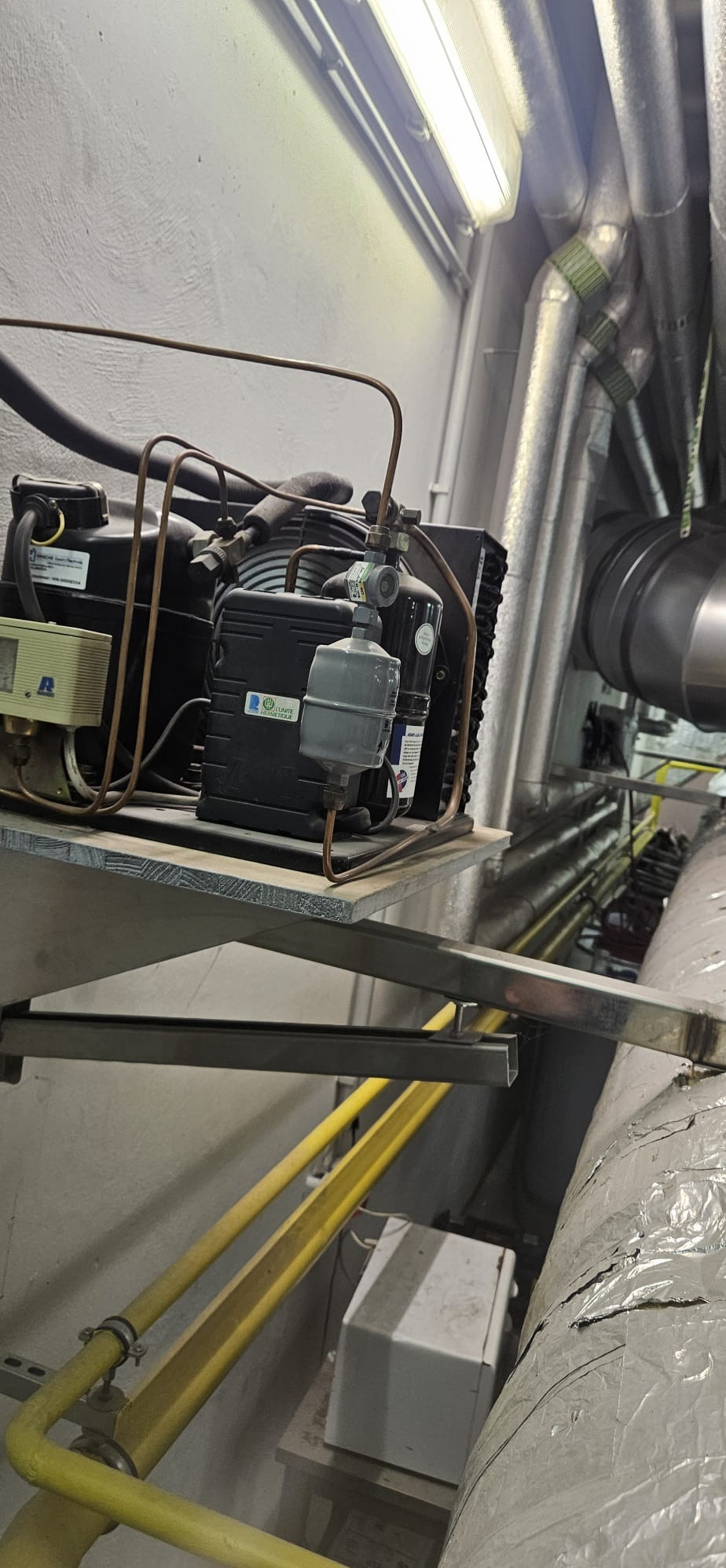 Zamontowany kompresor klimatyzacji na metalowej półce w pomieszczeniu technicznym, widoczne rury i instalacje. Szare ściany i sufit.