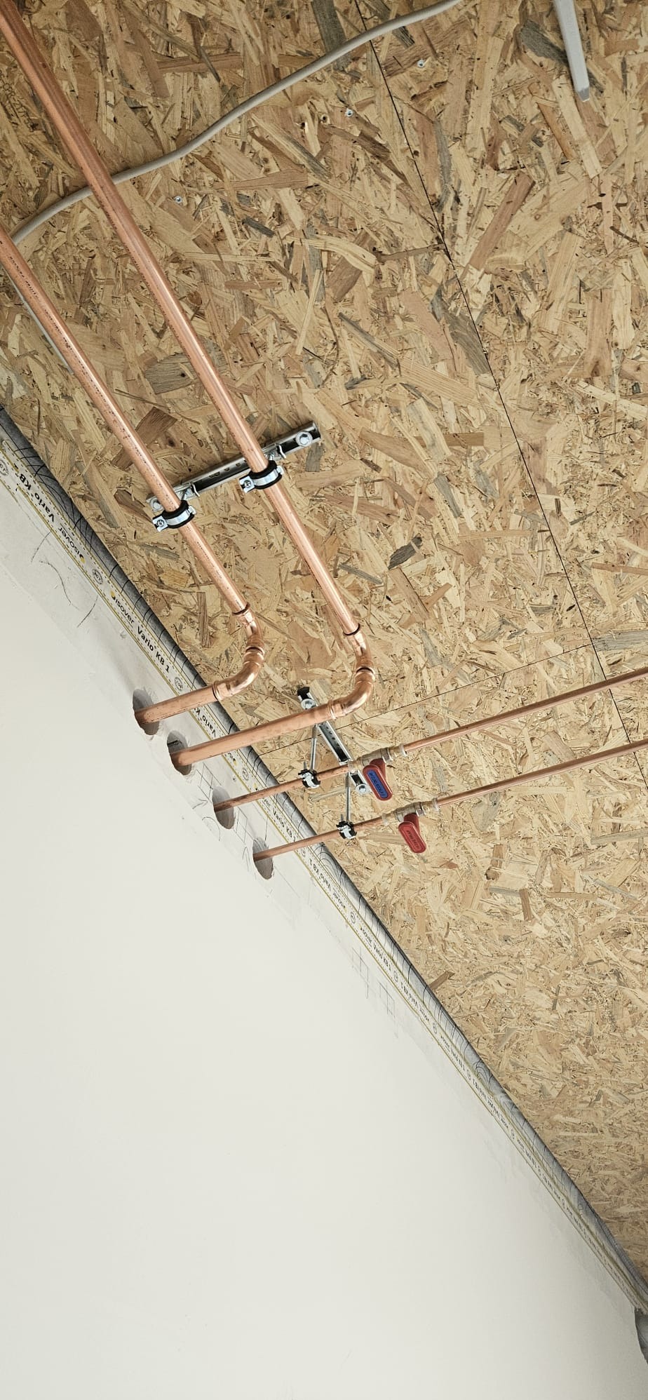 Miedziane rury instalacji wodnej zamocowane do sufitu z płyty OSB, widoczne zawory odcinające. Instalacja w trakcie montażu, sufit z widoczną izolacją.