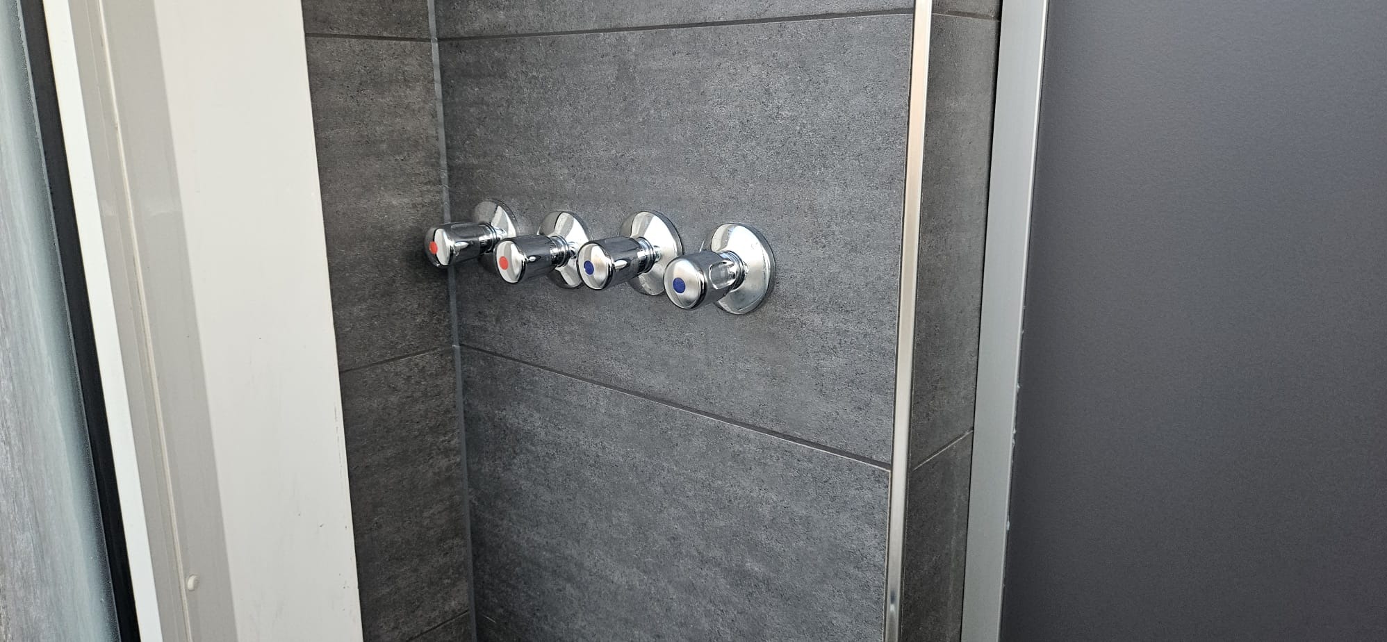 Cztery chromowane zawory z oznaczeniami ciepłej i zimnej wody, zamontowane na szarej, płytkowej ścianie w kabinie prysznicowej z aluminiowymi profilami.