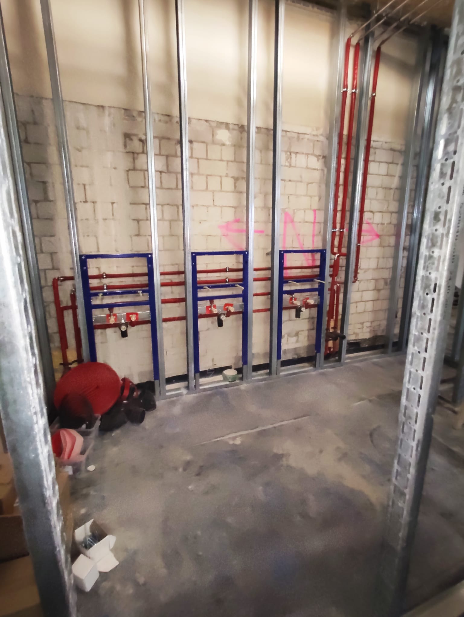 Wnętrze w trakcie instalacji hydraulicznej: widoczne rury (czerwone) i niebieskie ramy mocujące, surowe ściany z cegły i metalowe profile konstrukcyjne.