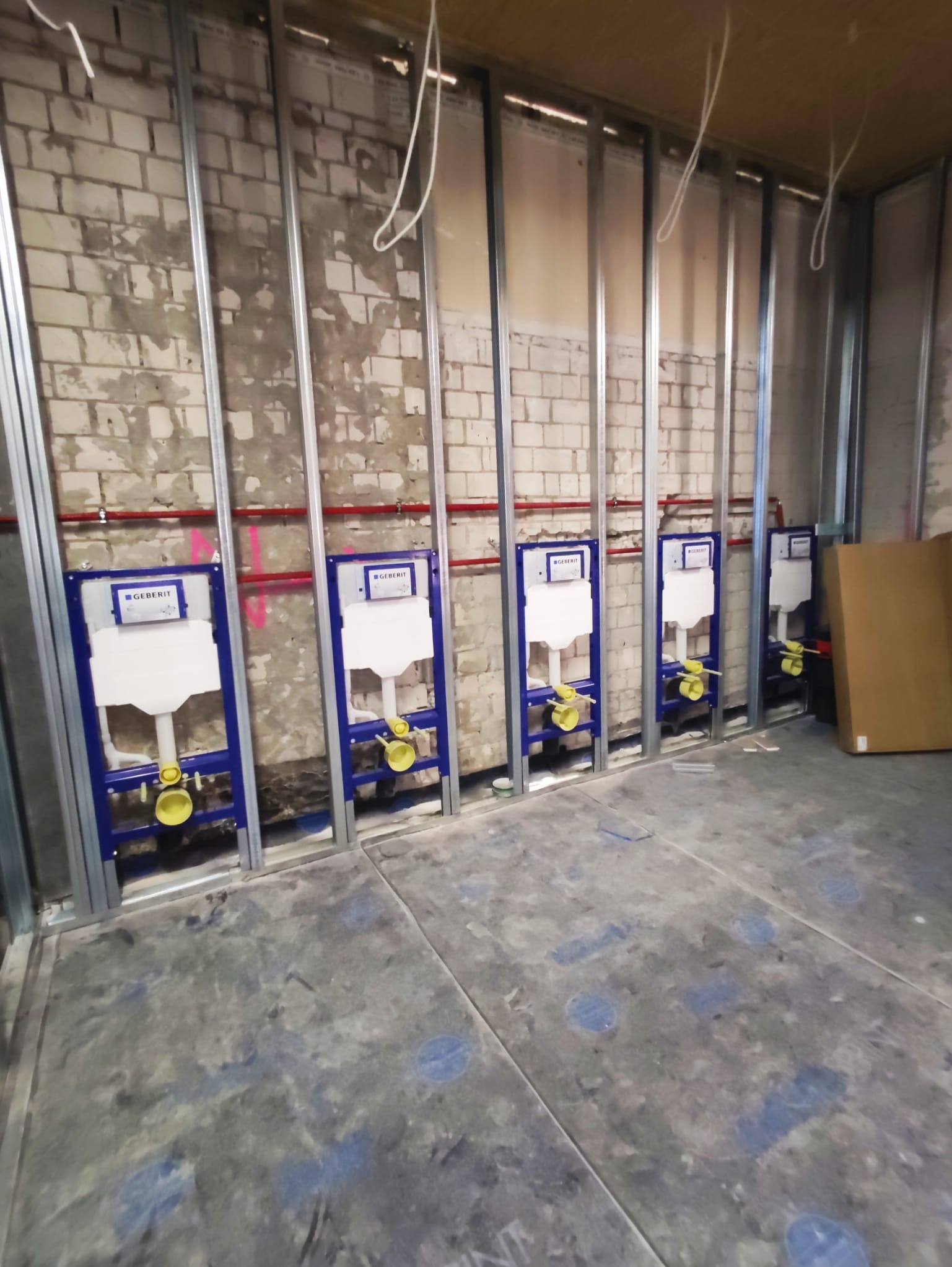 Rząd stelaży podtynkowych Geberit zamontowanych na ścianie z cegły, widoczne metalowe profile i instalacje. Surowy stan wykończenia wnętrza.