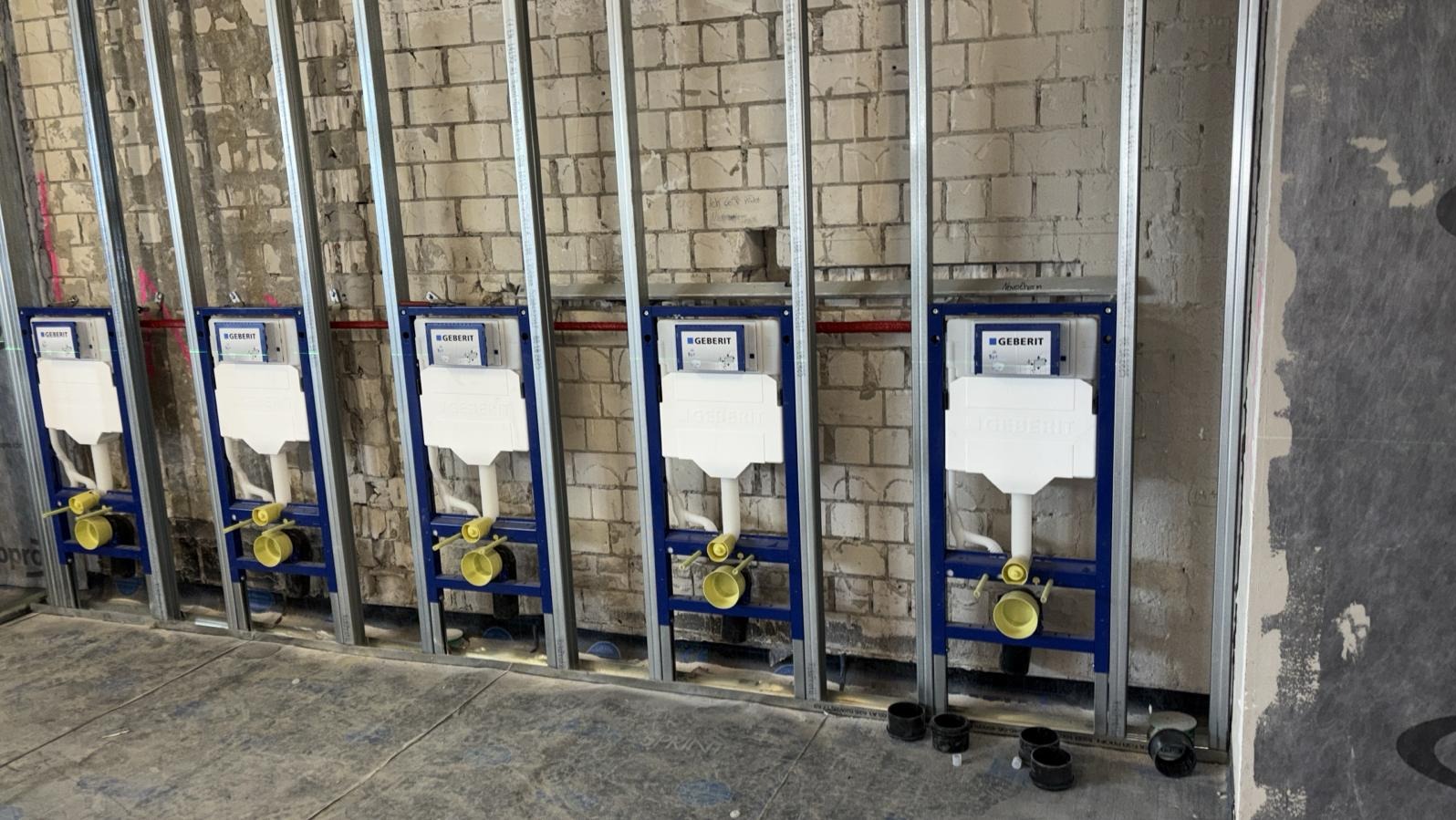 Cztery stelaże podtynkowe Geberit zamontowane w metalowej konstrukcji na tle surowej, ceglanej ściany. Widoczne elementy instalacji hydraulicznej i kanalizacyjnej.