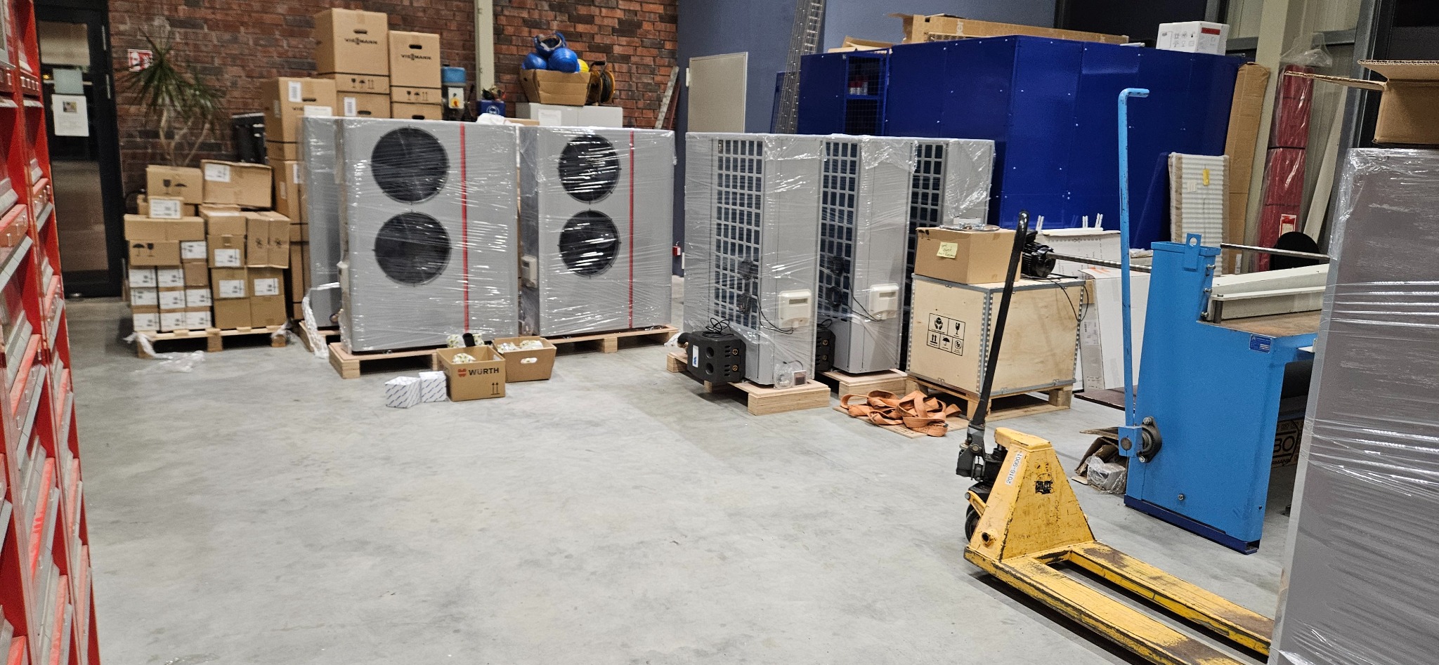 Klimatyzatory Viessmann, zapakowane w folię, stoją na paletach w magazynie. W tle widoczne kartony i niebieska obudowa urządzenia. Żółty wózek paletowy z przodu.