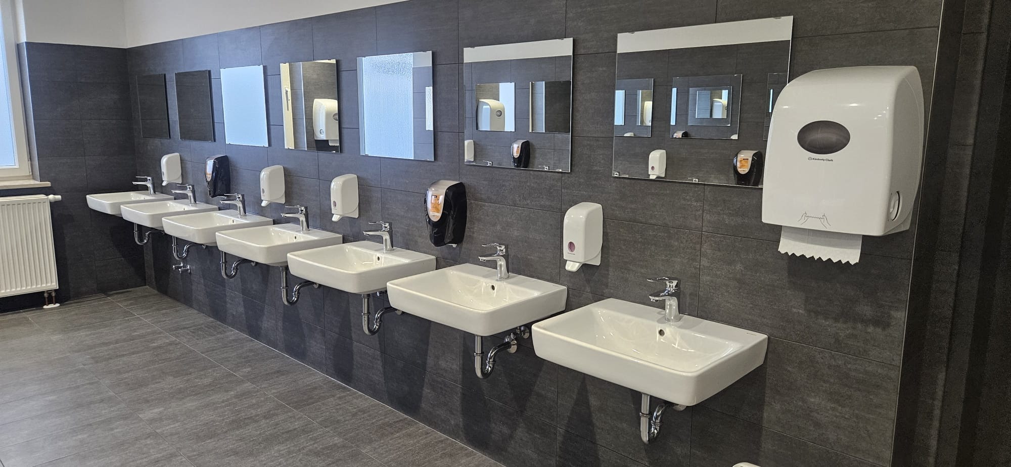 Rząd białych umywalek z chromowanymi kranami zamontowanych na szarej, kafelkowanej ścianie w publicznej toalecie. Nad umywalkami wiszą lustra i dozowniki mydła.