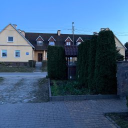 Jakub Jarzembowski - Fasada domu z numerem 21, z przodu dwa wysokie, gęste, zielone tuje, niewielki ogródek z kwiatami i studnia z brązowym daszkiem, kostka brukowa na podjeździe.