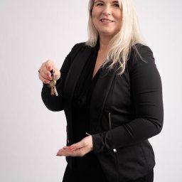 Własny Kąt - NIERUCHOMOŚCI I INWESTYCJE - ALEKSANDRA CZYKIETA - Uśmiechnięta kobieta w czarnym żakiecie i sukience, trzymająca w jednej dłoni pęk kluczy, a drugą dłoń wyciągniętą jakby coś oferowała, na jasnym tle.