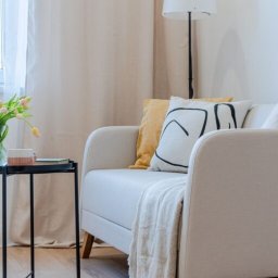 Własny Kąt - NIERUCHOMOŚCI I INWESTYCJE - ALEKSANDRA CZYKIETA - Jasny salon z beżową sofą, poduszkami w kolorze żółtym i z czarnym wzorem, stolikiem z tulipanami i lampą podłogową.