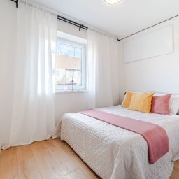 Własny Kąt - NIERUCHOMOŚCI I INWESTYCJE - ALEKSANDRA CZYKIETA - Jasna sypialnia z dużym oknem, białymi zasłonami, drewnianą podłogą i łóżkiem z żółtymi i różowymi poduszkami oraz narzutą w odcieniach bieli i różu.