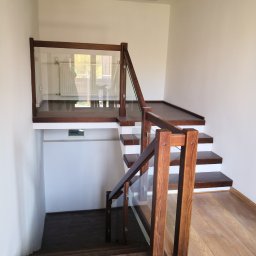 Własny Kąt - NIERUCHOMOŚCI I INWESTYCJE - ALEKSANDRA CZYKIETA - Dwukondygnacyjne schody z drewnianymi stopniami i balustradą ze szkła, widok z dołu na górny poziom z kaloryferem za szybą.