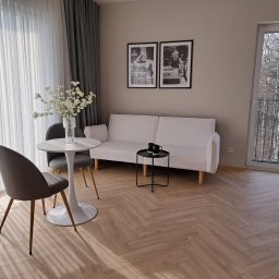 Własny Kąt - NIERUCHOMOŚCI I INWESTYCJE - ALEKSANDRA CZYKIETA - Jasne wnętrze pokoju z parkietem w jodełkę, białym stolikiem kawowym z kwiatami, szarą sofą, czarnym stolikiem pomocniczym z dwoma filiżankami i plakatami w czarnych ramkach na ścianie.