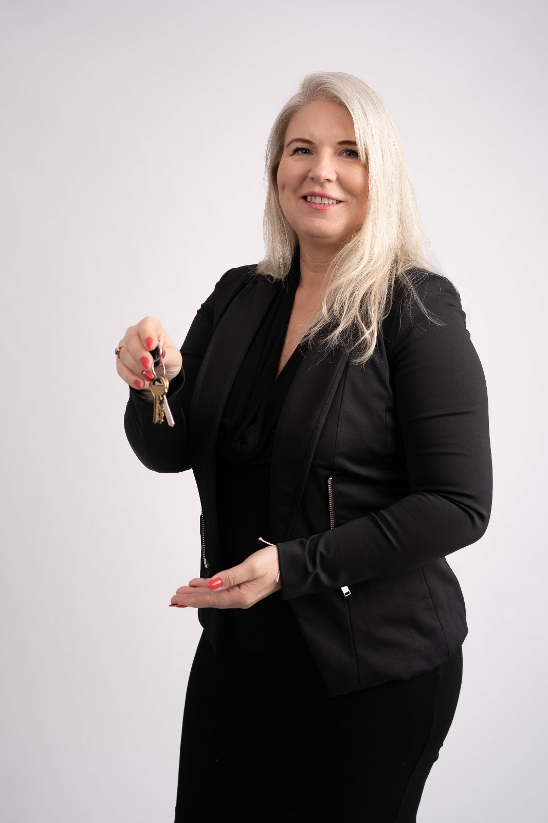 Uśmiechnięta kobieta w czarnym żakiecie i sukience, trzymająca w jednej dłoni pęk kluczy, a drugą dłoń wyciągniętą jakby coś oferowała, na jasnym tle.