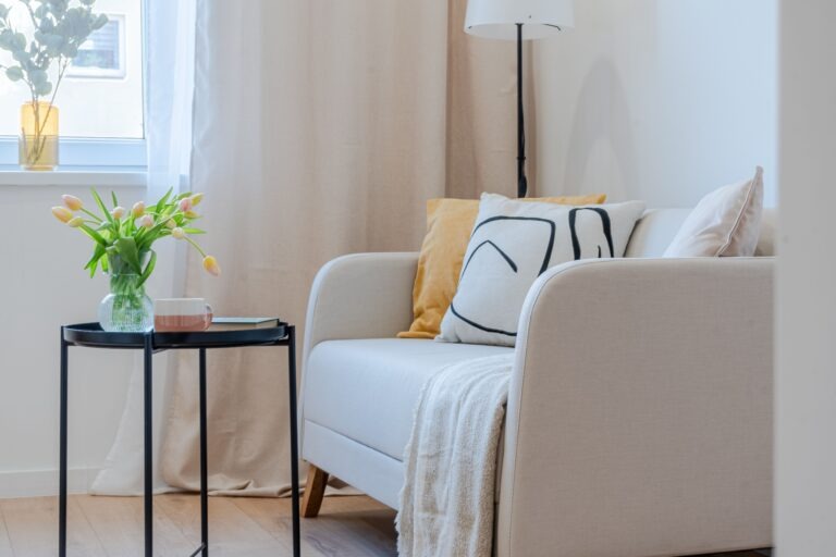 Jasny salon z beżową sofą, poduszkami w kolorze żółtym i z czarnym wzorem, stolikiem z tulipanami i lampą podłogową.