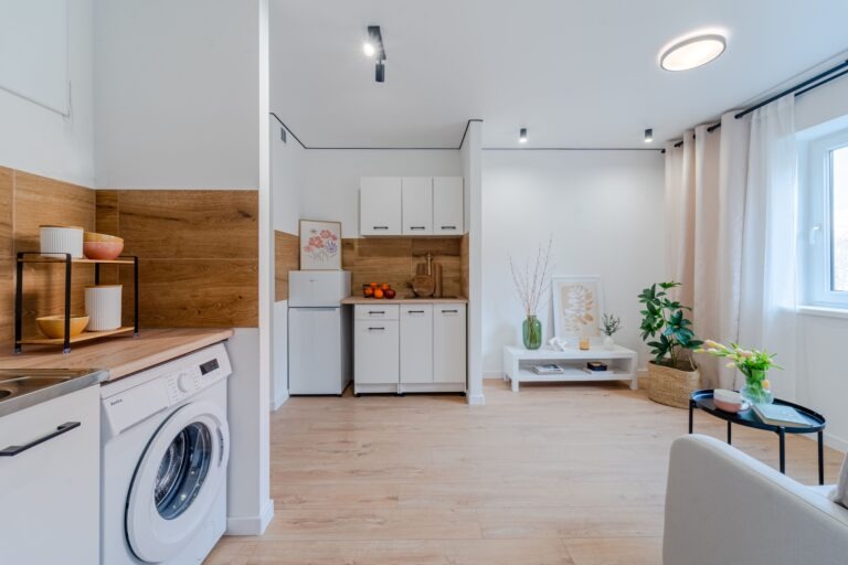 Jasne, nowocześnie urządzone studio z pralką w aneksie kuchennym, drewnianymi akcentami i minimalistycznym wystrojem.