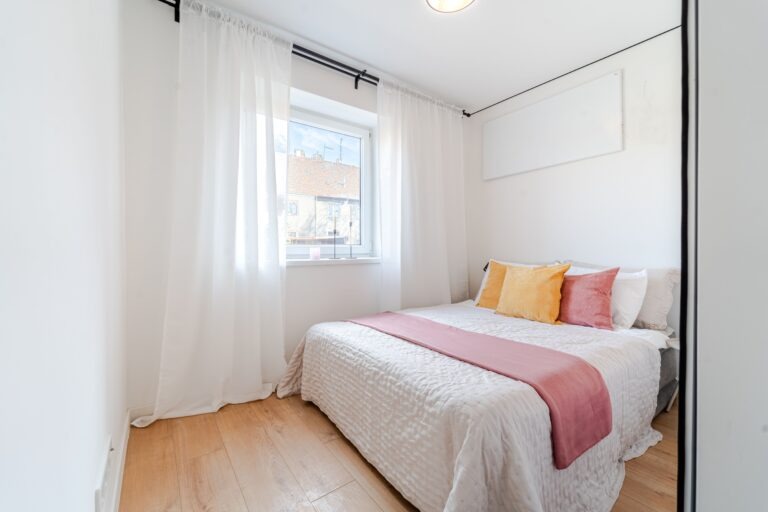 Jasna sypialnia z dużym oknem, białymi zasłonami, drewnianą podłogą i łóżkiem z żółtymi i różowymi poduszkami oraz narzutą w odcieniach bieli i różu.