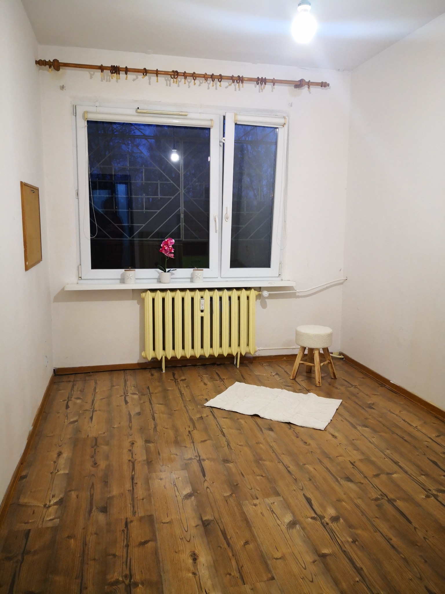 Pusta, jasna kawalerka w Rybniku z drewnianą podłogą, żółtym grzejnikiem pod oknem, białym dywanikiem i stołkiem.