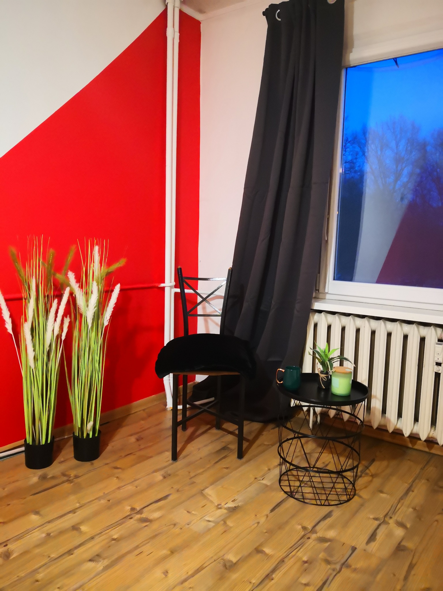 Wnętrze pokoju z drewnianą podłogą, ścianą w kolorze czerwono-białym, czarnym krzesłem z aksamitnym siedziskiem, stolikiem z roślinami i świecą oraz czarnymi zasłonami przy oknie.