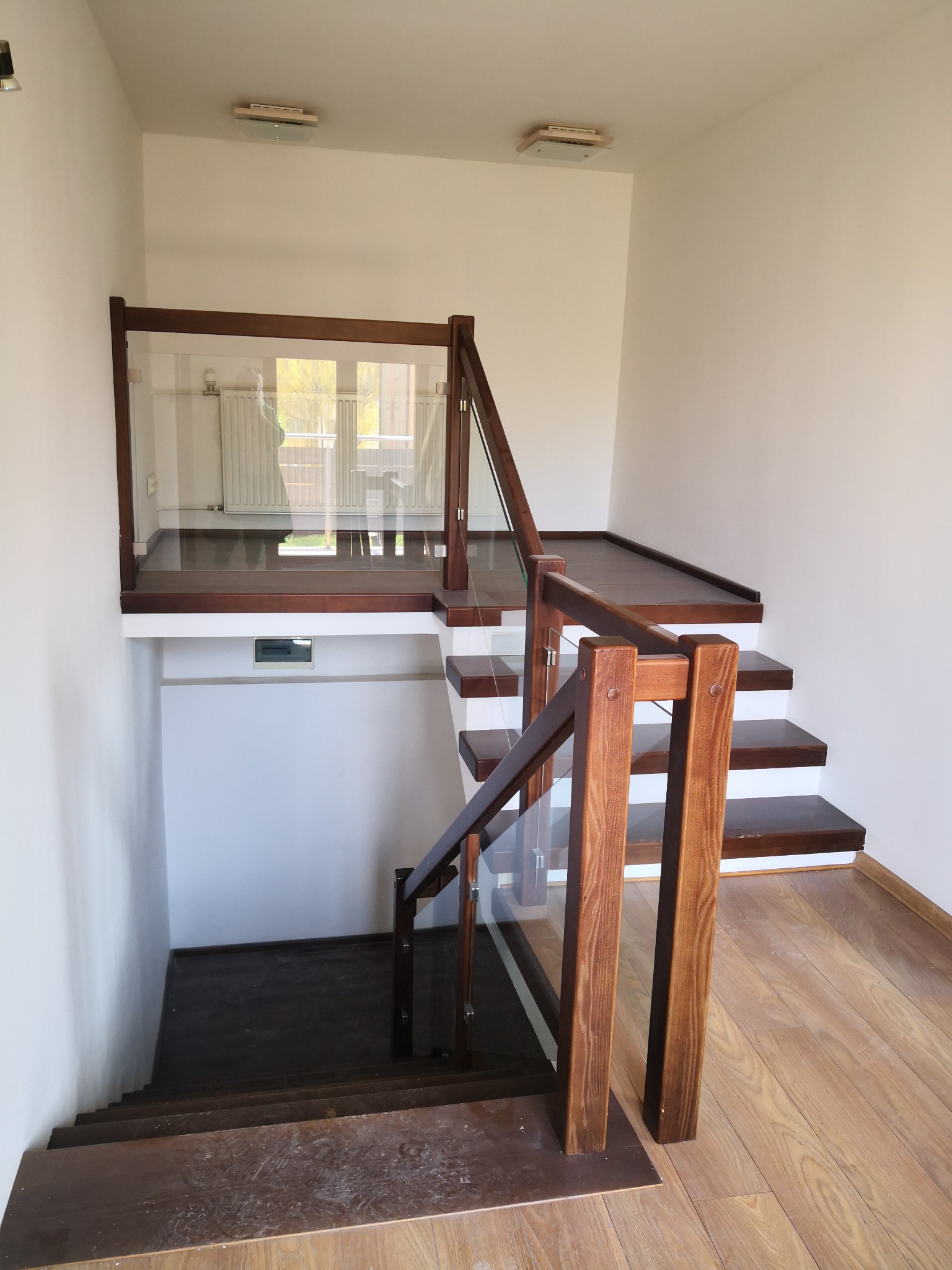 Dwukondygnacyjne schody z drewnianymi stopniami i balustradą ze szkła, widok z dołu na górny poziom z kaloryferem za szybą.