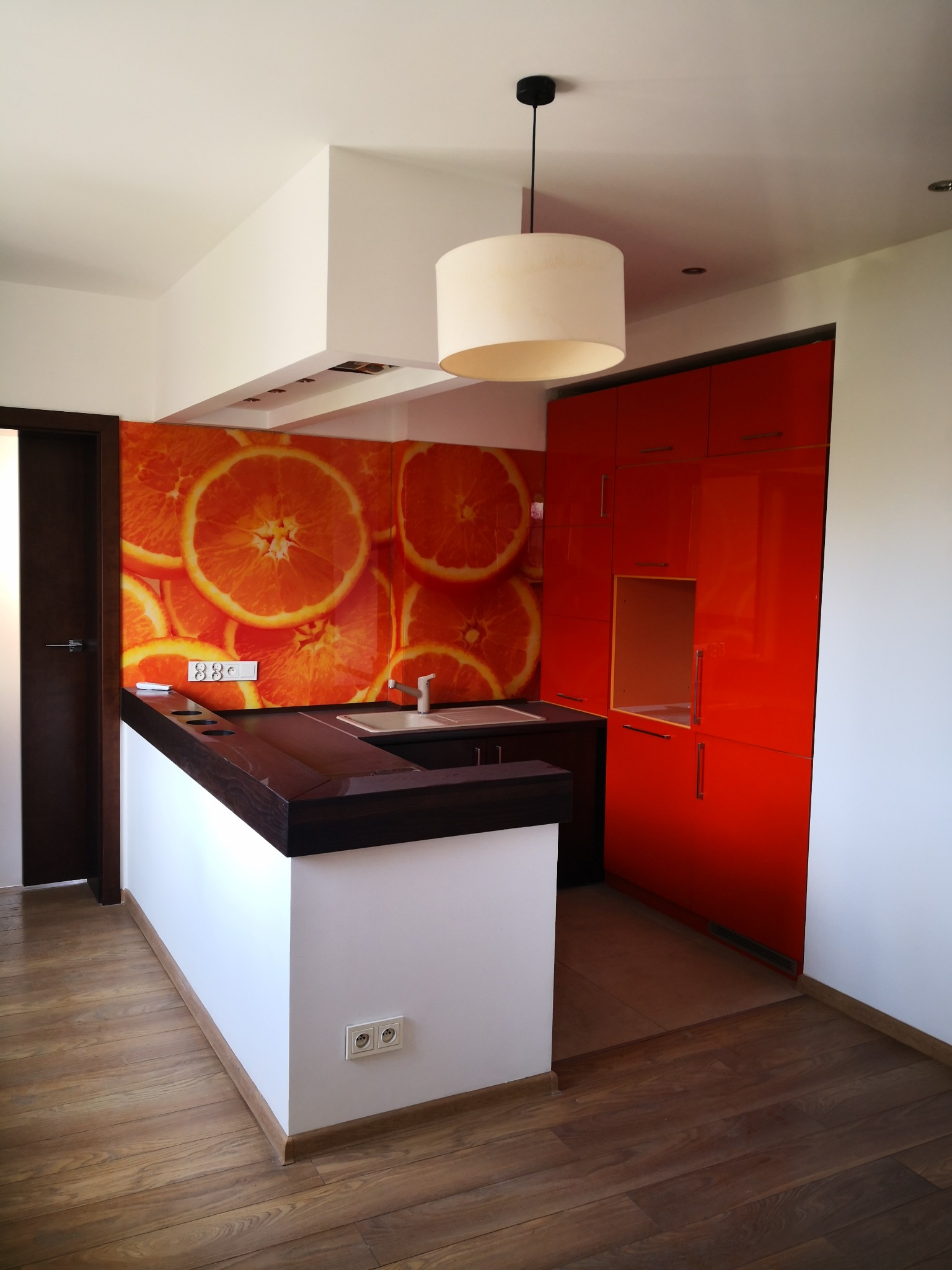 Nowoczesna kuchnia z pomarańczowymi akcentami: fototapeta z motywem pomarańczy, czerwone szafki na wysoki połysk, ciemnobrązowy blat i drewniana podłoga.
