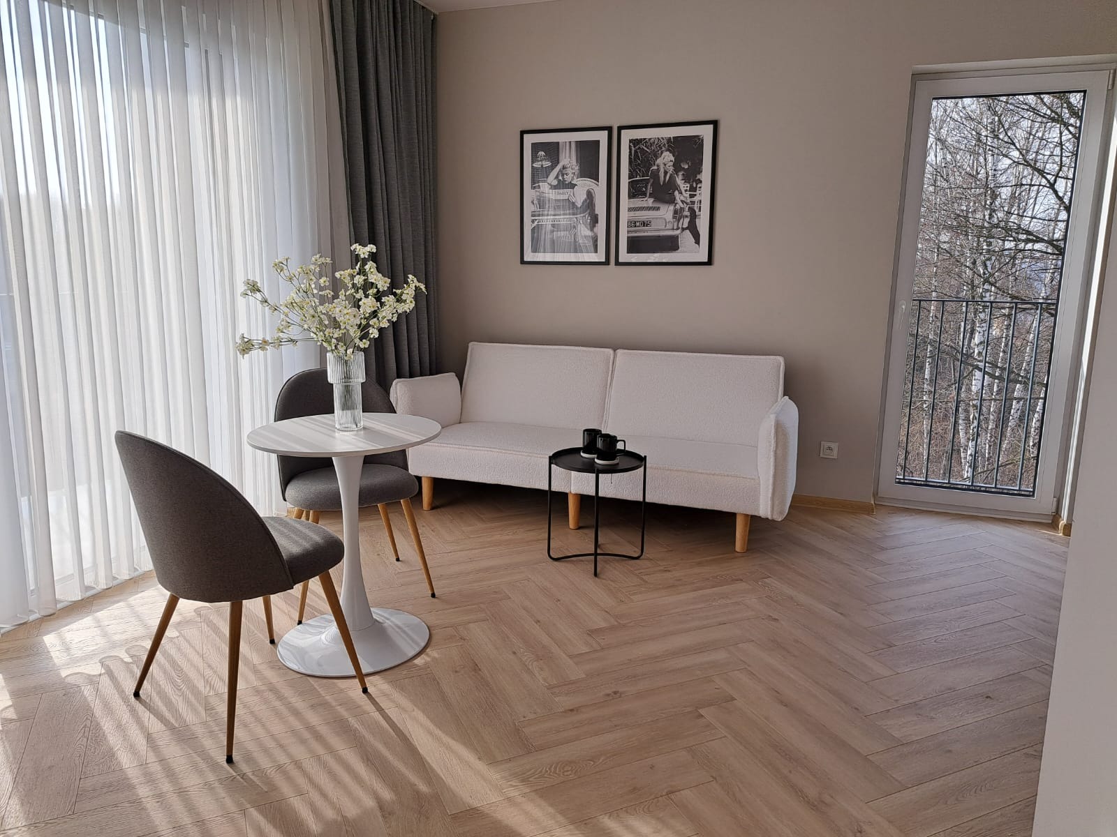 Jasne wnętrze pokoju z parkietem w jodełkę, białym stolikiem kawowym z kwiatami, szarą sofą, czarnym stolikiem pomocniczym z dwoma filiżankami i plakatami w czarnych ramkach na ścianie.