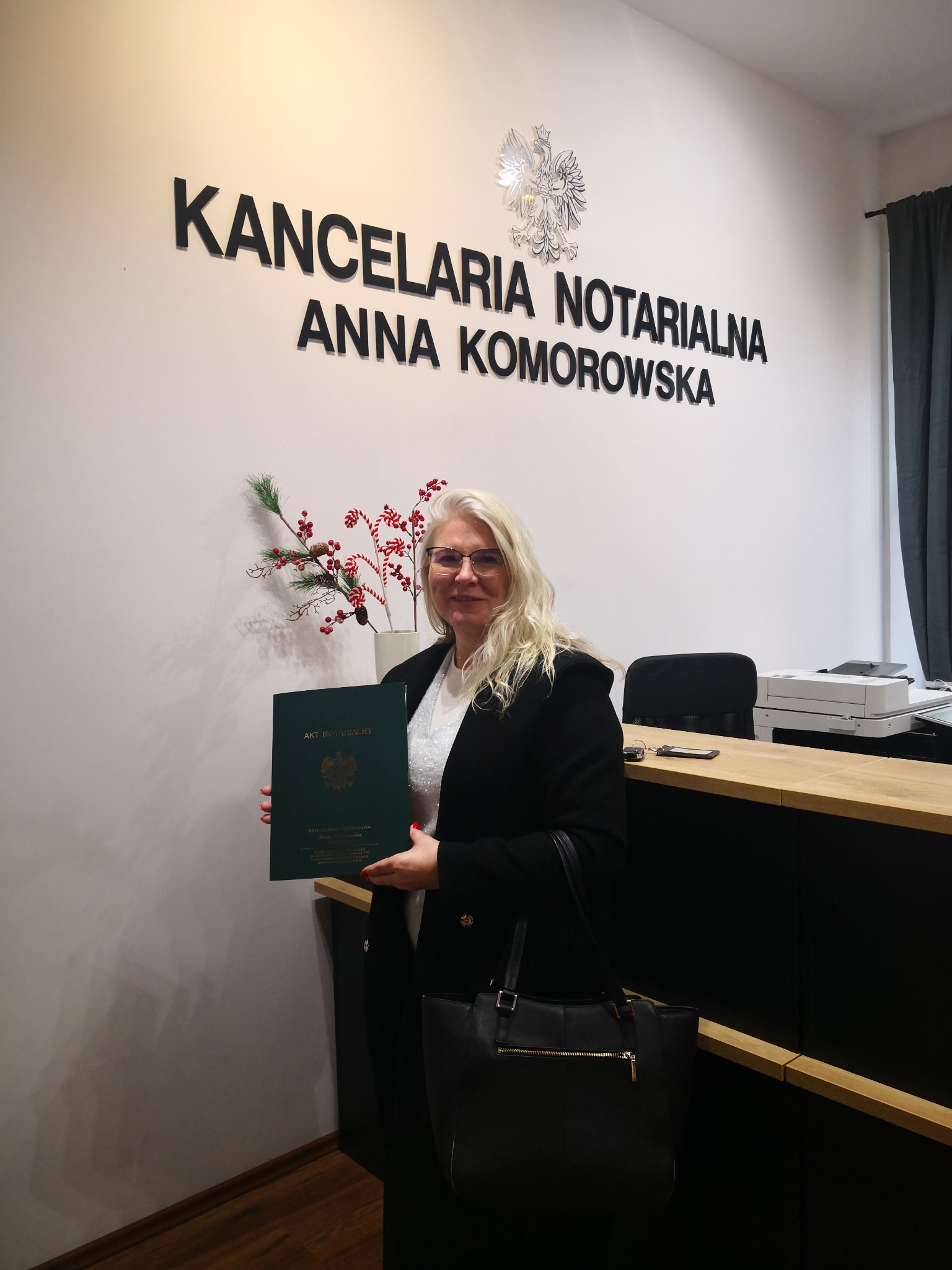 Kobieta w okularach trzyma akt notarialny na tle ściany z napisem 'Kancelaria Notarialna Anna Komorowska' i godłem Polski.