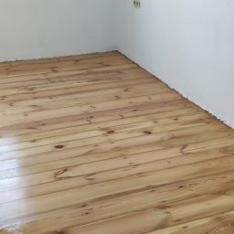 Parkiety Rafał Nowicki - Świeżo polakierowana podłoga z jasnego drewna w rogu pokoju, widoczne gniazdka elektryczne na białej ścianie, naturalne światło z okna.