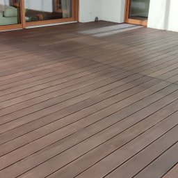 Parkiety Rafał Nowicki - Nowa, brązowa podłoga z desek na tarasie, widoczne przesuwne drzwi balkonowe i dekoracyjne lampki wiszące.