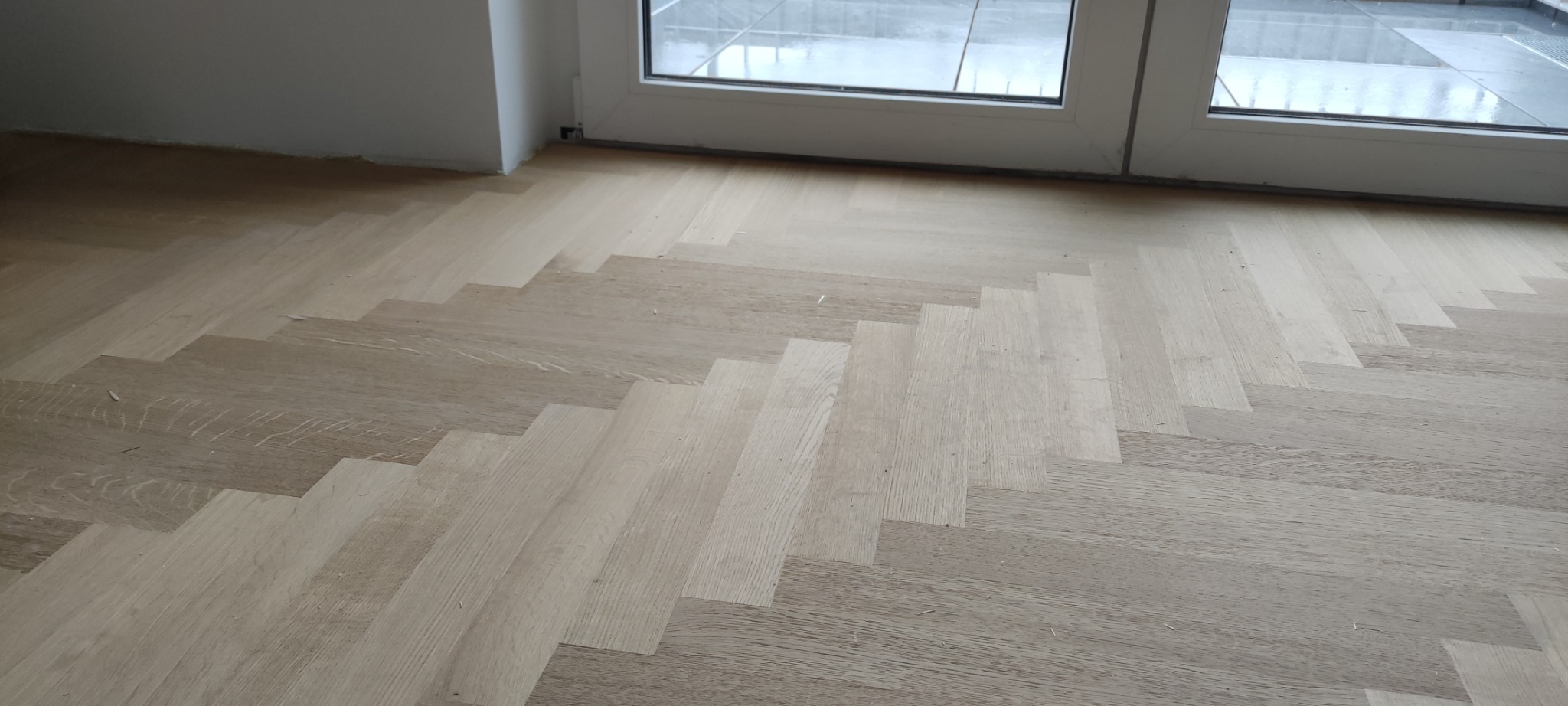 Jasny parkiet ułożony we wzór jodełki francuskiej, widoczne drobne resztki po montażu, przy białych drzwiach balkonowych.