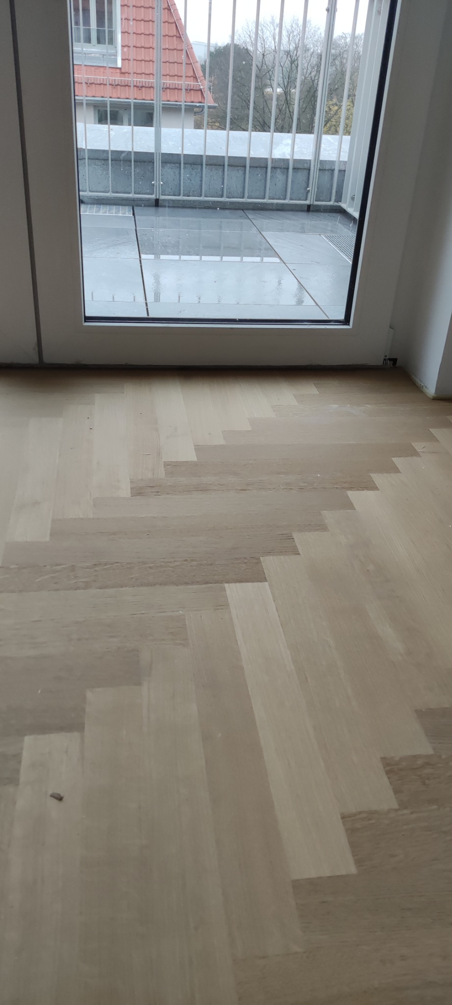 Jasny parkiet ułożony we wzór jodełki francuskiej, widok z niskiej perspektywy w kierunku okna balkonowego z metalową barierką.