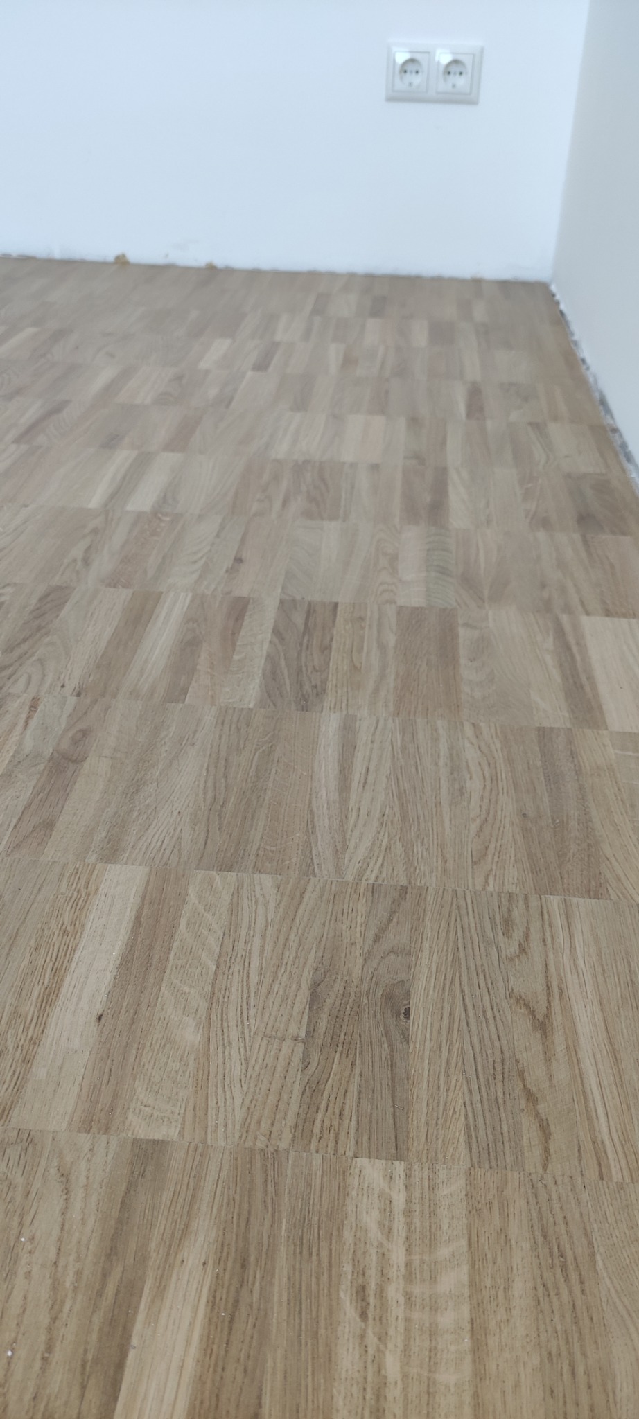 Ułożony parkiet w jodełkę francuską, widoczne gniazdka elektryczne na białej ścianie i minimalne listwy przypodłogowe.
