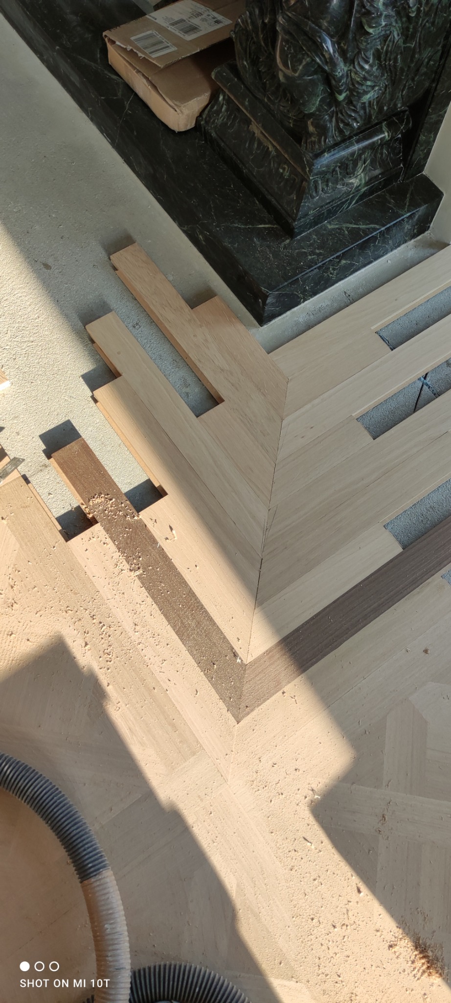 Fragment układanego parkietu w jodełkę z ciemniejszymi wstawkami, widoczne niedocięte elementy i pył po obróbce, obok marmurowy fragment kominka.