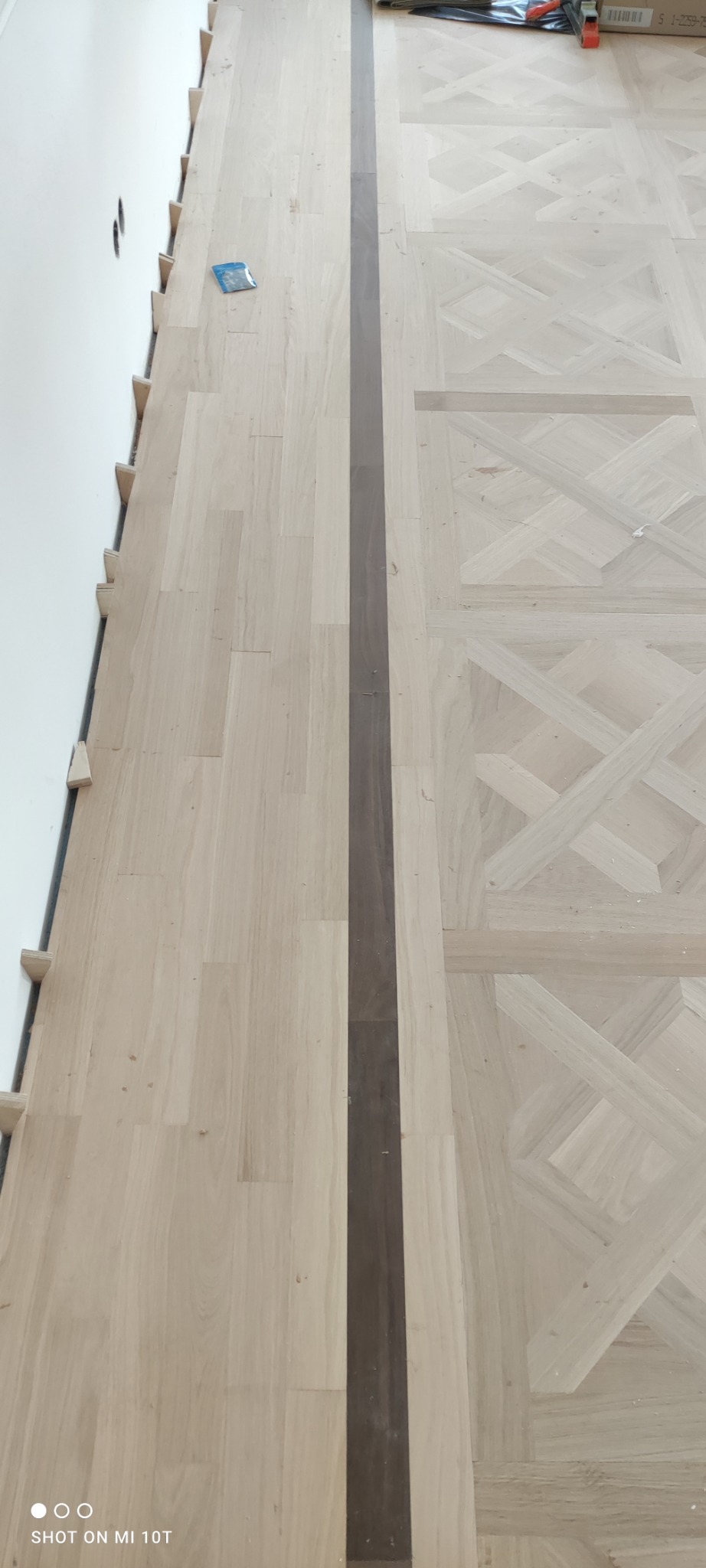 W trakcie montażu parkietu z ciemną intarsją, widoczne kliny dystansowe przy ścianie i fragmenty narzędzi.