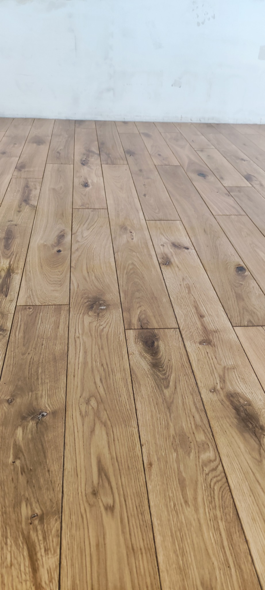 Ułożona podłoga z paneli drewnianych w odcieniach brązu, widoczne sęki i naturalne usłojenie drewna, przy białej ścianie z widocznymi śladami po malowaniu.