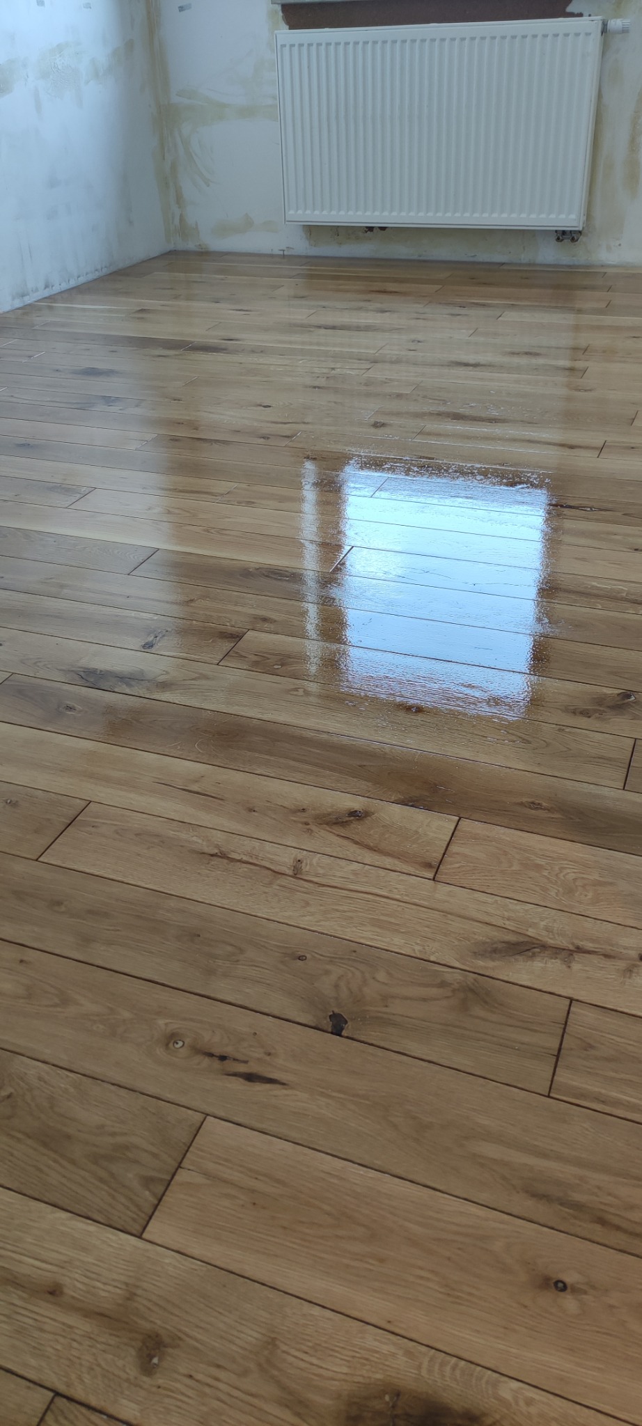 Lśniąca, nowo położona drewniana podłoga parkietowa z widocznym odbiciem okna i grzejnikiem na ścianie w tle.