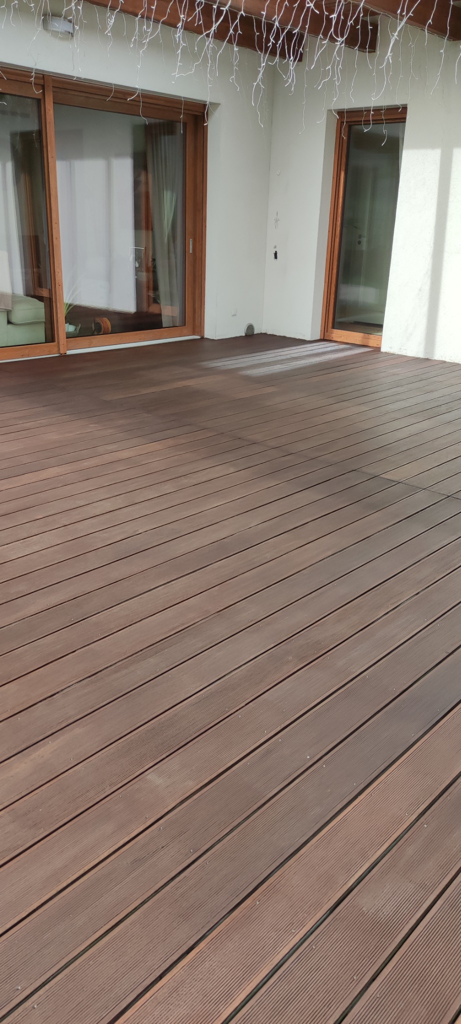 Nowa, brązowa podłoga z desek na tarasie, widoczne przesuwne drzwi balkonowe i dekoracyjne lampki wiszące.