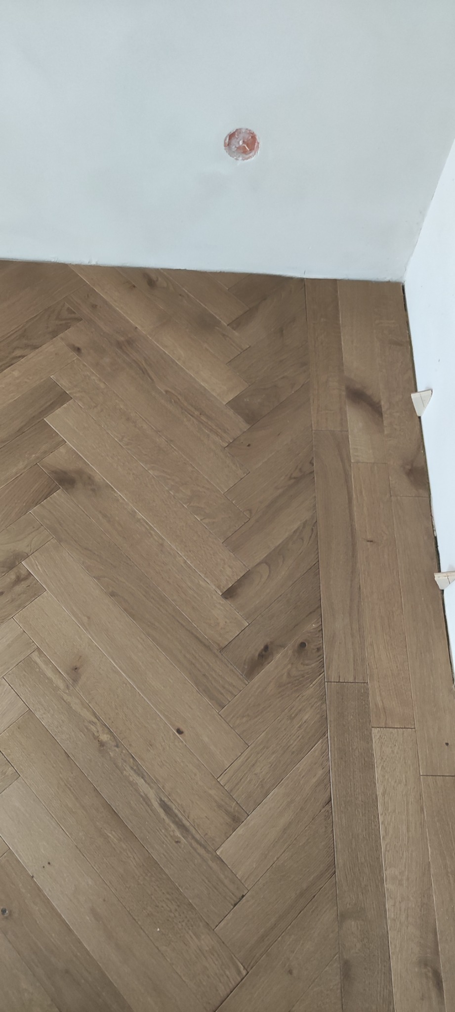 Parkiet ułożony we wzór jodełki, jasne drewno, widoczne listwy dystansowe przy ścianie, surowy stan wykończenia, gniazdko elektryczne wystające ze ściany.