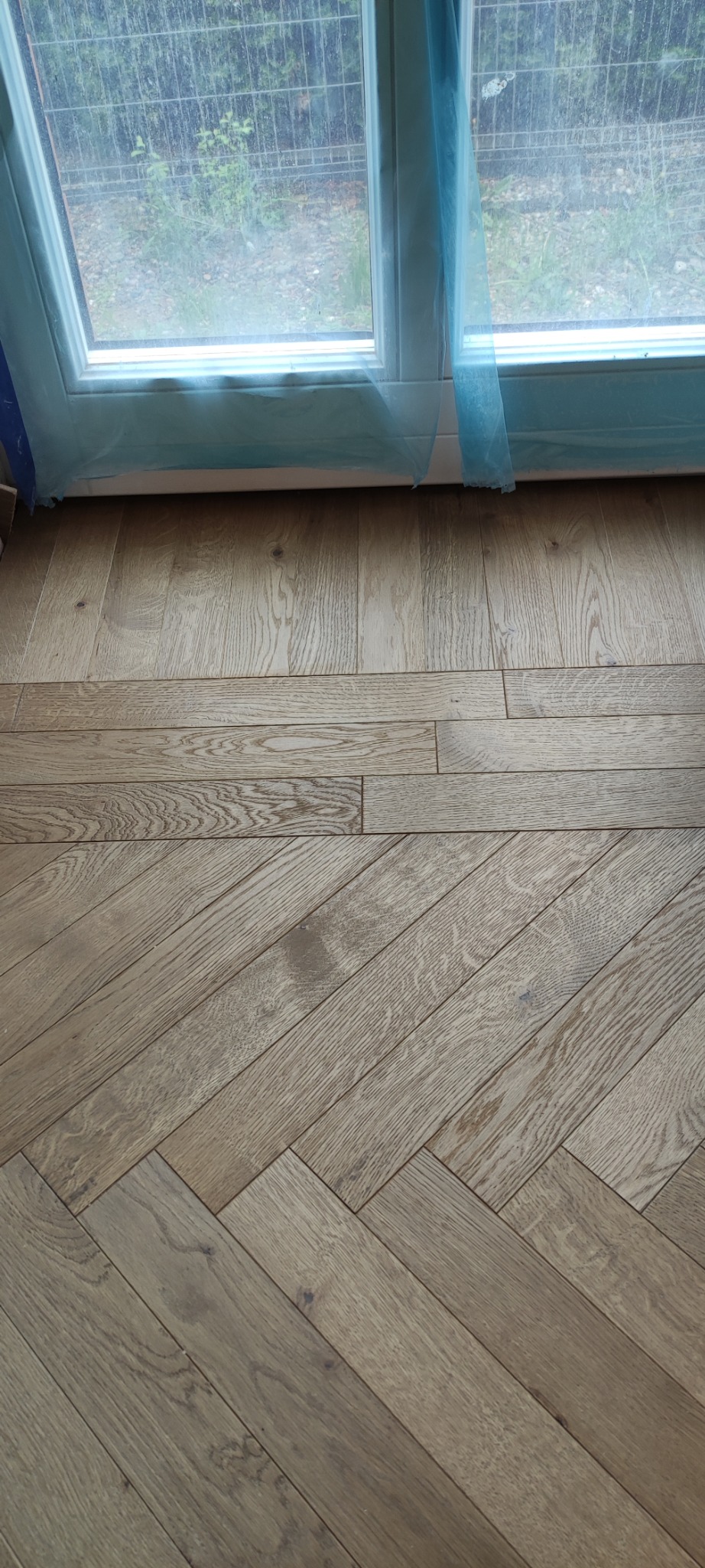 Parkiet ułożony w jodełkę francuską, widoczny przy oknie zabezpieczonym folią malarską. Jasne drewno, naturalny odcień. Fragment podłogi.