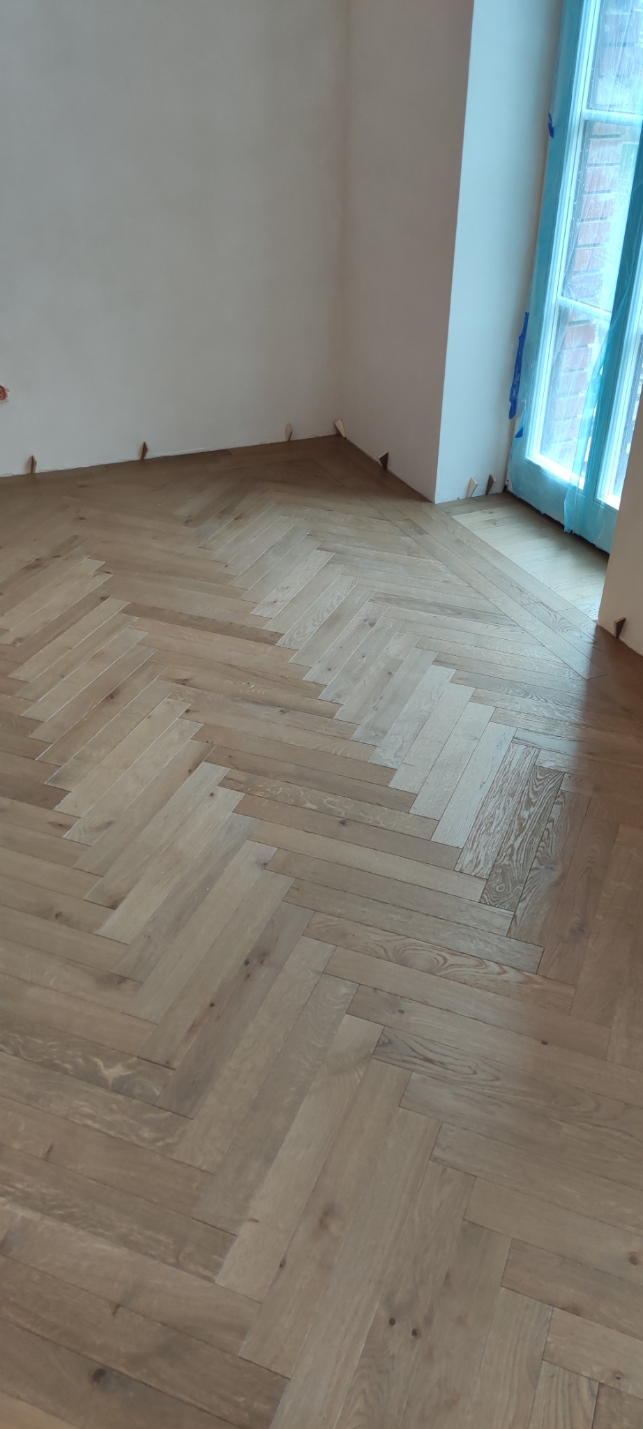 Parkiet ułożony w jodełkę francuską w jasnym odcieniu drewna, zabezpieczony folią okno. Listwy przypodłogowe jeszcze nie zamontowane. Widok na fragment podłogi w pokoju.