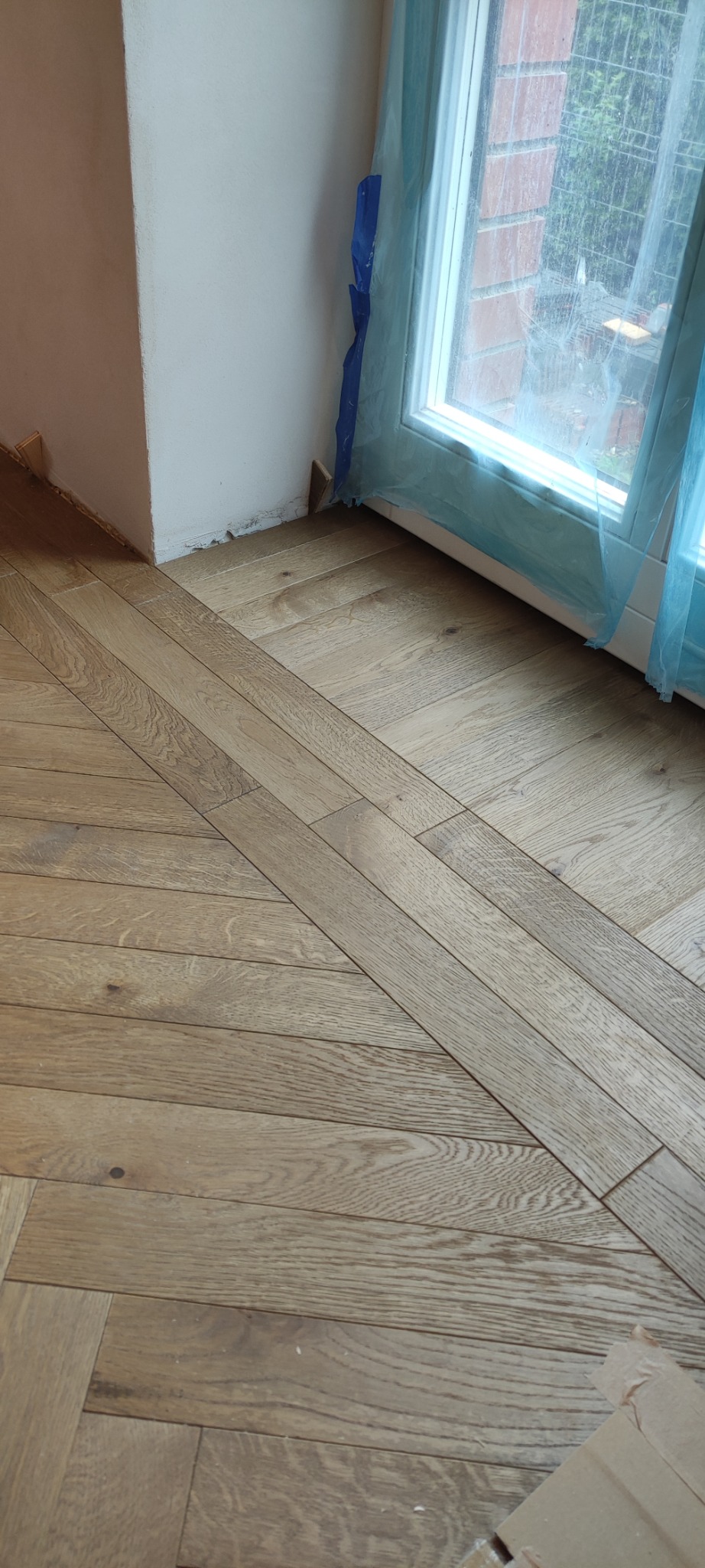 Ułożony parkiet w jodełkę francuską, jasne drewno, widoczne zabezpieczenie okna folią malarską. Detal wykończenia podłogi przy ścianie i oknie.