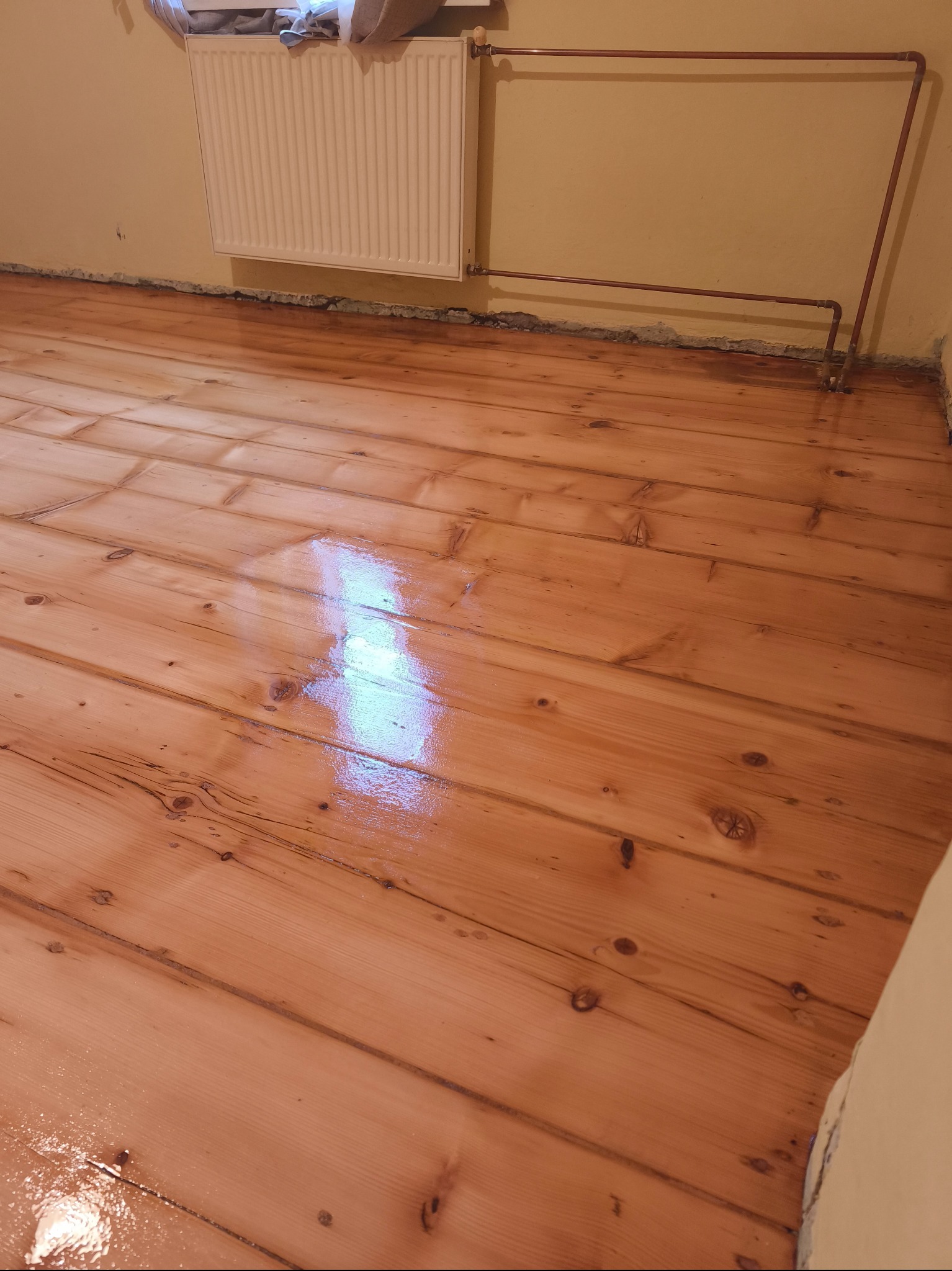 Lakierowana podłoga z desek po cyklinowaniu, widoczne ślady lakieru, ściana z grzejnikiem i rurami. Podłoga w odcieniach brązu i beżu, świeżo po renowacji.