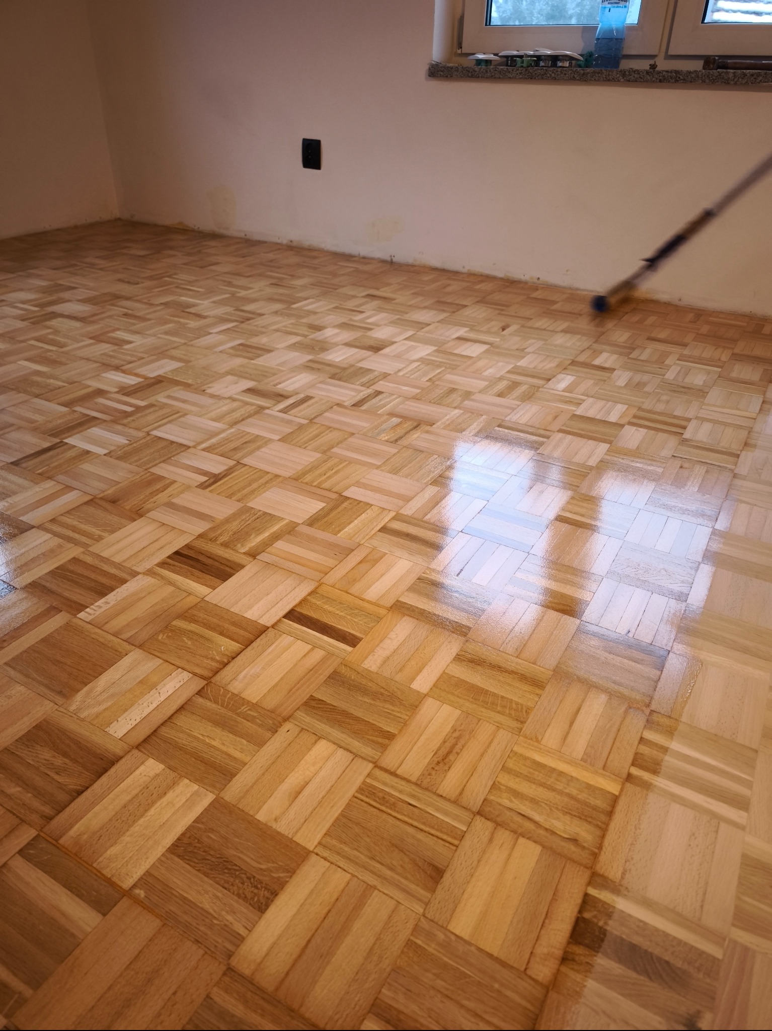 Lakierowany parkiet w jodełkę podczas prac wykończeniowych. Widoczny wałek do lakierowania i refleksy światła na świeżo położonej warstwie lakieru.