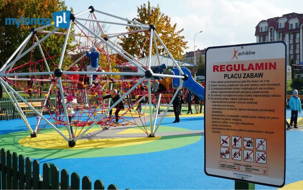 Kolorowy plac zabaw z konstrukcją linową, dzieci wspinają się. Tablica z regulaminem po prawej, budynek w tle. Jasne, słoneczne oświetlenie.