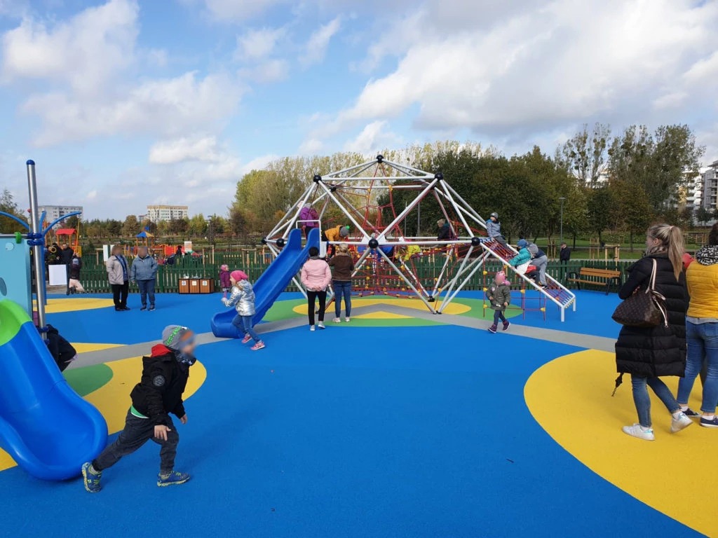 Kolorowy plac zabaw z konstrukcją w kształcie kuli, zjeżdżalnią i dziećmi bawiącymi się na tle drzew i zabudowań miejskich w słoneczny dzień.